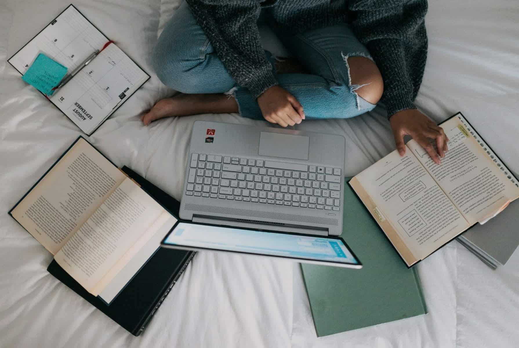 Student sitting on bed studying with books and laptop